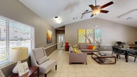 Located at 4235 SE 10th Pl in Gainesville, Florida, this single-floor community room offers a welcoming and functional open-plan layout designed for gathering and relaxation. The space is anchored by soaring vaulted ceilings with a ceiling fan, creating an airy, expansive feel throughout. Light-toned ceramic tile flooring runs seamlessly across the entire floor, complementing the warm taupe two-tone walls accented by white chair rail molding.

The living area features a large sectional sofa adorned with colorful accent pillows, flanked by accent chairs and a coffee table, creating a comfortable lounge zone. Vibrant wall art adds personality and color to the space. A separate seating nook near the windows provides a quieter retreat, bathed in natural light filtering through plantation-shuttered windows.

The kitchenette area is equipped with light wood cabinetry, a refrigerator, microwave, and ample counter space, making it ideal for small gatherings. Adjacent dining tables with chairs offer flexible seating arrangements. A bookshelf and television station provide entertainment and reading options. French doors with direct exterior access invite natural light and connect the interior to the outdoors, enhancing the overall openness of this well-appointed community space.