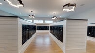 This dedicated mailroom is designed to serve a large residential community with efficiency and style. The space is organized around a central island unit and perimeter wall-mounted mailbox banks, all finished in sleek black metal with individual keyed compartments to accommodate a high volume of residents. The mailbox enclosures are framed by crisp white shiplap-style paneling, creating a clean contrast against the black hardware and contributing to the room's modern aesthetic.

Light hardwood-style flooring runs throughout the space, enhancing its bright and open feel. Natural light filters in through a row of clerestory windows positioned near the ceiling line, supplemented by recessed lighting and striking ring-shaped pendant fixtures with dark bronze frames that hang overhead, providing ample illumination throughout the room.

The walls are finished in a soft neutral gray, and a colorful piece of abstract artwork is displayed on one accent wall clad in dark shiplap paneling, adding a touch of personality to the functional space. A built-in shelf below the artwork provides a convenient surface for sorting mail. The overall layout allows for easy resident circulation, and the thoughtful design elevates what is typically a purely utilitarian space into a polished, well-finished amenity area.