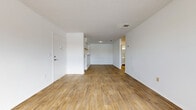 This single-floor unit offers a clean, functional layout with a consistent design language throughout. The main living area is open and spacious, featuring warm-toned wood-look flooring that extends across the entire unit. Crisp white walls and a textured ceiling provide a neutral backdrop, while a built-in shelving nook near the kitchen pass-through adds a practical architectural detail. The front entry opens directly into this expansive living space, which flows naturally toward additional rooms.

The kitchen is efficiently designed with white cabinetry, white countertops, and white appliances including a full-size refrigerator and electric range. An under-cabinet ventilation hood and ample upper and lower storage make the space fully equipped for everyday use. The kitchen connects to the living area via a half-wall pass-through, maintaining an open feel.

The bedroom is generously proportioned, featuring the same wood-look flooring, bright white walls, and a window fitted with blinds that allows natural light to fill the room. A secondary space visible in the images features a glazed French door and lighter flooring, offering flexibility as a dining area or additional room.

The bathroom is neatly finished with a white vanity and single-basin sink, a large mirror, and a combined tub and shower unit with chrome grab bars and a sliding glass door enclosure. Wood-look flooring continues here, tying the unit together with a cohesive, well-maintained aesthetic.