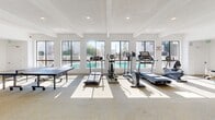 Located at 1549 Placentia Ave in Newport Beach, CA, this generously sized single-floor community amenity space offers a well-rounded recreational experience designed to serve a variety of activities and interests. The open layout is unified by neutral carpet flooring, white walls, and a textured ceiling with exposed beams, creating a clean and bright atmosphere throughout.

The fitness area is positioned along a wall of large windows that overlook an outdoor pool, flooding the space with natural light. It is equipped with multiple treadmills, elliptical machines, a multi-use weight bench, free weights, kettlebells, and a full-length mirror — providing a comprehensive workout environment.

Adjacent to the fitness zone is a dedicated game and recreation area featuring both a ping-pong table and a pool table, offering casual entertainment options for residents.

At the far end of the space, a comfortable lounge area serves as a social hub, anchored by a striking floor-to-ceiling brick fireplace adorned with a large decorative clock. Seating arrangements with sofas and coffee tables invite relaxation, while a dining area nearby adds functional versatility. Windows lining the perimeter of the lounge allow for ample natural light and an open, airy feel. Recessed lighting throughout ensures the entire space is well-lit at any time of day.