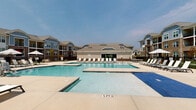 Located at 1025 Hirschfeld Dr in Fayetteville, NC, this well-appointed community amenity center serves as a central gathering hub for residents. The interior showcases contemporary design with light wood-style flooring, recessed lighting, and a neutral color palette that creates a bright, welcoming atmosphere throughout.

The lounge area is anchored by a striking floor-to-ceiling stacked stone fireplace with a mounted TV, flanked by built-in shelving and complemented by a circular statement chandelier. Comfortable seating arrangements and a game table make this space ideal for socializing and relaxation.

The clubhouse kitchen features sleek dark cabinetry topped with white quartz countertops, a bold herringbone-patterned tile accent wall, and a generous island with bar-height seating — a perfect setup for community gatherings and events. Large sliding glass doors connect the interior seamlessly to the outdoor amenity deck, offering direct views of the resort-style pool.

Outside, the expansive pool area includes multiple swimming zones, a shallow wading section, and a sun shelf, all surrounded by a spacious deck lined with lounge chairs, umbrellas, and seating. The gated community exterior features multi-story residential buildings with brick and board-and-batten facades, manicured landscaping, and a dedicated outdoor recreation area.