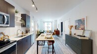 This thoughtfully designed single-floor residence offers a seamless open-concept layout that connects the kitchen, dining, and living areas into one cohesive, light-filled space. The kitchen is outfitted with sleek navy-blue cabinetry, stainless steel appliances, a white countertop island with bar seating, and track lighting overhead — creating a functional yet contemporary cooking environment. Light wood-tone flooring runs throughout, lending warmth and continuity across every room.

The living area is generously proportioned, anchored by a large window that frames natural light and views of the surrounding streetscape. The space flows comfortably from the dining island toward a relaxed seating area, making it well-suited for both entertaining and everyday living.

The residence includes two bedrooms, each with its own distinct character. One features crisp white walls, light-filtering curtains, and a serene, minimal aesthetic. The second bedroom makes a bold design statement with a dramatic floral-patterned accent wall paired with a tufted upholstered headboard, adding depth and personality to the space.

Two full bathrooms complete the layout — one featuring white subway tile surrounding a full soaking tub and shower combination, and the other offering a clean vanity with access to a walk-in closet. The overall ambiance is modern, bright, and welcoming throughout.