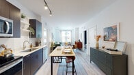 This thoughtfully designed single-floor residence offers a seamless open-concept layout that connects the kitchen, dining, and living areas into one cohesive, light-filled space. The kitchen is outfitted with sleek navy-blue cabinetry, stainless steel appliances, a white countertop island with bar seating, and track lighting overhead — creating a functional yet contemporary cooking environment. Light wood-tone flooring runs throughout, lending warmth and continuity across every room.

The living area is generously proportioned, anchored by a large window that frames natural light and views of the surrounding streetscape. The space flows comfortably from the dining island toward a relaxed seating area, making it well-suited for both entertaining and everyday living.

The residence includes two bedrooms, each with its own distinct character. One features crisp white walls, light-filtering curtains, and a serene, minimal aesthetic. The second bedroom makes a bold design statement with a dramatic floral-patterned accent wall paired with a tufted upholstered headboard, adding depth and personality to the space.

Two full bathrooms complete the layout — one featuring white subway tile surrounding a full soaking tub and shower combination, and the other offering a clean vanity with access to a walk-in closet. The overall ambiance is modern, bright, and welcoming throughout.