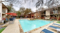 Located along Preston Road in the north Dallas area of Texas, this two-story residential community combines classic brick and neutral-toned exterior architecture with well-maintained communal outdoor spaces. The buildings feature traditional pitched rooflines, prominent chimneys, and private balconies that overlook the central courtyard.

At the heart of the community is a generously sized outdoor swimming pool, framed by natural stone coping and surrounded by a spacious pool deck. The pool area is well-appointed with lounge chairs, dining-height seating, and vibrant red umbrella shade structures, providing ample space for both relaxation and socializing. Patches of lush green turf are integrated into the poolside design, adding a refreshing visual contrast to the stone surfaces.

The grounds are enriched by mature oak trees that provide natural canopy and shade throughout the courtyard. Carefully curated landscaping features decorative grasses, agave plants, flowering shrubs, and raised stone planters, giving the outdoor spaces a polished, resort-like atmosphere. Dedicated lounge areas with chaise seating are thoughtfully positioned throughout the green spaces, offering quiet retreats away from the main pool.

The two-floor building layout ensures convenient access to both ground-level and upper-level units, each benefiting from direct views of the landscaped courtyard and pool area.