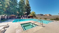 Located at 275 East Minnesota Avenue in Turlock, California, this outdoor amenity area centers around a generously sized swimming pool with clear, blue-green water and clearly marked depth gradients along the pool deck. The single-level layout is thoughtfully designed to maximize comfort and usability, with a dedicated spa/hot tub positioned adjacent to the main pool for easy access.

The surrounding pool deck provides ample space for relaxation, furnished with a row of chaise lounges ideal for sunbathing, as well as shaded seating areas outfitted with patio tables, chairs, and cantilever umbrellas in warm orange tones. Steps and handrails offer safe, convenient entry into the pool at multiple points.

The space is framed by tall, mature trees — including towering pines — that provide natural shade and privacy, creating a tranquil retreat within the community. A low perimeter wall encloses the area, with neatly maintained low-lying shrubs and landscaping lining its base. Adjacent buildings feature a Mediterranean-influenced architectural style with warm brick tones and red clay tile roofing, complementing the overall character of the outdoor space.

Under the abundant California sunshine, this pool area offers a well-appointed, resort-style outdoor environment suited for both active recreation and leisurely relaxation.