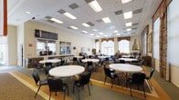 This single-floor community space is designed to accommodate group gatherings, dining, and social activities in a welcoming and functional environment. The main hall is expansive, featuring numerous round white tables paired with black chairs, arranged to encourage conversation and community interaction. A drop ceiling fitted with recessed rectangular fluorescent lighting ensures the space is well-lit throughout.

Large windows dressed with decorative valances allow natural light to filter into the room, creating a warm and inviting atmosphere. A standout architectural feature is the stone fireplace situated along one wall, adding a cozy and traditional character to the otherwise practical layout. The flooring transitions between a neutral vinyl border and a patterned carpet in the central area, helping to define the gathering space.

Along one side of the room, a service or activity counter with a large pass-through window suggests the presence of a kitchen or resource area, ideal for serving meals or distributing materials. Informational boards and displays are mounted on the walls, indicating the space may serve an educational or community service function. Overall, the hall offers a comfortable, multi-purpose environment suited for dining, events, classes, or community programs.
