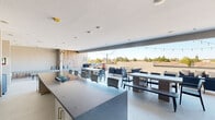 Located along North Scottsdale Road in Scottsdale, Arizona, this rooftop patio is a well-appointed outdoor amenity space designed for socializing and relaxation. The layout seamlessly blends a partially covered indoor-outdoor zone with an open-air terrace, creating a versatile environment suited to the Arizona climate.

The covered section features a sleek granite-topped outdoor kitchen bar equipped with a stainless steel sink, under-counter refrigeration, and a mounted flat-screen television — ideal for entertaining. Recessed ceiling lighting and wall-mounted fans ensure comfort throughout the year. Adjacent to the kitchen, a warmly accented wood-panel wall hosts built-in bench seating with patterned cushions, offering a cozy lounge nook.

The open patio extends outward with multiple seating configurations, including long communal dining tables with chairs and benches, as well as modular lounge furniture arranged around a central gathering area. String lights strung overhead add an inviting ambiance for evening gatherings.

Panoramic views of the Scottsdale rooftops, desert landscaping, and distant mountain silhouettes — including what appears to be Camelback Mountain — are visible from the terrace. A dedicated grill station and additional outdoor lounge clusters round out this dynamic and well-equipped rooftop retreat.