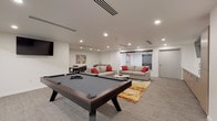 Located at 612 W Main Ave in downtown Spokane, Washington, this contemporary amenity lounge is thoughtfully designed to serve as a social and recreational hub. The single-level space features a clean, modern aesthetic defined by crisp white walls, recessed lighting, and neutral carpet flooring that creates a bright and welcoming atmosphere throughout.

At the center of the room, a full-size pool table with a sleek dark finish serves as the focal point of the recreational area. Flanking this game zone is a wall-mounted entertainment setup featuring multiple flat-screen TVs alongside built-in cabinetry with light wood finishes and white quartz countertops, providing both style and functional storage.

The lounge seating area is furnished with a pair of generously sized gray sofas accented with bold red throw pillows, arranged around a modern marble-top coffee table and anchored by a vibrant abstract area rug in warm gold, red, and blue tones. This arrangement encourages comfortable conversation and relaxed gathering. A separate dining nook with a round table and chairs offers an additional space for more structured socializing.

The space also includes a glass-enclosed area visible from the main lounge, adding a sense of depth and transparency to the overall layout. The combination of recreational amenities, comfortable furnishings, and modern finishes makes this lounge a well-appointed destination for community engagement.