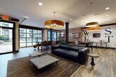 The Club Room at 820 W Marietta St NW is a well-appointed communal space designed in an industrial-chic aesthetic that blends raw architectural elements with refined furnishings. The open single-floor layout is unified by warm wood-plank flooring that flows throughout, complemented by exposed brick accent walls and bold black structural columns that reinforce the industrial character of the space.

Overhead, large copper-toned drum pendant lights serve as striking focal points, casting warm ambient light across the room. Recessed lighting supplements the natural light that pours in through expansive floor-to-ceiling steel-framed windows and glass garage-style doors, offering views of the outdoor surroundings and creating a bright, airy atmosphere.

The lounge area anchors the room with a substantial black leather sectional sofa, concrete-top coffee tables, and layered area rugs, arranged to encourage comfortable gathering. Railroad-themed artwork and decorative gear motifs displayed throughout speak to the building's industrial heritage.

A fully equipped wet bar features rich wood cabinetry, granite countertops, a stainless steel sink, and undercounter appliances set against an exposed brick backsplash. A dining and work table with bar-height seating provides additional functionality. A flat-screen television and a foosball table round out the entertainment offerings, making the space ideal for social events and casual recreation.