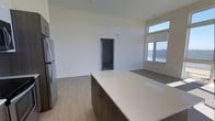 This single-floor space showcases a clean, contemporary design with an open-concept layout that seamlessly connects the kitchen and living areas. The kitchen is a standout feature, centered around a generous island with a crisp white quartz countertop and dark cabinetry with sleek hardware, offering both ample prep space and a modern aesthetic. Stainless steel appliances, including a full-size refrigerator, complete the well-appointed kitchen. Light wood-toned flooring runs throughout the space, complementing the bright white walls and contributing to the airy, modern feel. Elevated ceilings enhance the sense of openness, while large windows along one wall flood the interior with natural light and frame impressive water views, bringing the outdoors in. A doorway leads to an additional room, extending the functional layout of the unit. The overall ambiance is fresh, minimalist, and move-in ready, with a neutral palette that allows for a variety of interior design styles. The combination of thoughtful finishes, natural light, and scenic views makes this a comfortable and visually appealing living space.