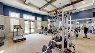 This fitness facility is thoughtfully designed to support a full range of workout activities across two dedicated exercise rooms and a living area. The main exercise room is anchored by striking architectural details, including coffered ceilings accented with rustic wood beams and recessed lighting, all set against bold navy blue upper walls and crisp white wainscoting below. The space is generously equipped with cardio machines including treadmills, ellipticals, a stair climber, and rowing machines, alongside a multi-station cable weight system, free weights, and a weight bench. Full-length mirrors line one wall, enhancing the sense of spaciousness, while mounted flat-screen televisions provide entertainment throughout. The second exercise room features soaring ceilings and an expansive wall of floor-to-ceiling windows that flood the space with natural light and offer open views of outdoor greenery and a pool area. This room is ideal for functional training, featuring stability balls, resistance bands, plyo boxes, and a stationary bike. Carpeted flooring runs throughout both rooms, and glass-paneled doors provide connectivity between spaces. The overall ambiance is bright, modern, and motivating, combining high-end finishes with a practical, well-organized layout suited for diverse fitness needs.