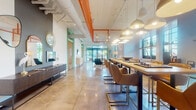 Located at 1017 Lake Ave in Lake Worth Beach, Florida, this single-floor space showcases a compelling blend of industrial and eclectic design. Polished concrete floors run throughout, reflecting light and tying together the open, expansive layout. Exposed ductwork and structural beams are painted white and accented with bold orange and green architectural details, lending an energetic, urban character to the interior.

The space is organized into several distinct zones. A long communal high-top table, flanked by tan and gold leather chairs and suspended dome pendant lights, serves as a central gathering and workspace hub. Floor-to-ceiling grid windows line the perimeter, flooding the interior with generous natural light and offering views of outdoor greenery.

Multiple lounge areas are furnished with cognac leather sectional sofas, rattan egg chairs, layered area rugs, and curated vinyl record wall displays, creating an inviting, lived-in atmosphere. A dedicated game area features a billiards table set against a textured OSB accent wall with a mounted flat-screen television. Decorative cacti, tropical plants, and eclectic artwork contribute to the relaxed, creative ambiance that defines this dynamic communal environment.