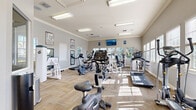 Located at 1222–1226 Floral Springs Blvd in Port Orange, Florida, this fitness center offers a comprehensive workout environment housed in a single-floor facility. The space is designed with functionality and comfort in mind, featuring neutral gray walls, crown molding, and durable carpet tile flooring laid in a herringbone-style pattern that adds a refined touch to the athletic setting.

Natural light pours in through multiple windows fitted with white plantation shutters, creating a bright and energizing atmosphere throughout the room. Overhead fluorescent lighting supplements the natural light, ensuring the space remains well-lit at all hours. A wall-mounted flat-screen television is positioned for easy viewing from various workout stations, adding an entertainment element to the exercise experience.

The equipment selection is extensive, including upright and recumbent stationary bikes, elliptical cross-trainers, treadmills, and a variety of strength-training machines targeting multiple muscle groups. Strategically placed mirrors along the walls enhance the sense of openness while allowing users to monitor their form. The layout provides ample circulation space between machines, promoting a safe and comfortable workout flow.

Additional amenities include a wall-mounted AED device, waste receptacles, and motivational fitness posters. The entry area features glass-panel doors, contributing to the open and welcoming character of the facility.
