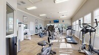Located at 1222–1226 Floral Springs Blvd in Port Orange, Florida, this fitness center offers a comprehensive workout environment housed in a single-floor facility. The space is designed with functionality and comfort in mind, featuring neutral gray walls, crown molding, and durable carpet tile flooring laid in a herringbone-style pattern that adds a refined touch to the athletic setting.

Natural light pours in through multiple windows fitted with white plantation shutters, creating a bright and energizing atmosphere throughout the room. Overhead fluorescent lighting supplements the natural light, ensuring the space remains well-lit at all hours. A wall-mounted flat-screen television is positioned for easy viewing from various workout stations, adding an entertainment element to the exercise experience.

The equipment selection is extensive, including upright and recumbent stationary bikes, elliptical cross-trainers, treadmills, and a variety of strength-training machines targeting multiple muscle groups. Strategically placed mirrors along the walls enhance the sense of openness while allowing users to monitor their form. The layout provides ample circulation space between machines, promoting a safe and comfortable workout flow.

Additional amenities include a wall-mounted AED device, waste receptacles, and motivational fitness posters. The entry area features glass-panel doors, contributing to the open and welcoming character of the facility.