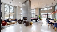 Located at 1629 S Razorback Rd in Fayetteville, Arkansas, this contemporary community lounge is a thoughtfully designed gathering space that blends modern architecture with local University of Arkansas spirit. The single-floor layout is anchored by dramatically high ceilings that amplify the sense of openness, while expansive floor-to-ceiling windows flood the interior with abundant natural light and offer views of the surrounding area.

A striking stone-clad double-sided column serves as the architectural centerpiece of the main lounge area, housing a sleek linear fireplace on one side and a wall-mounted flat-screen television on the other. The polished concrete flooring reflects light throughout and provides a clean, modern foundation for the eclectic furnishings. A full-sized billiards table with a bold red felt surface occupies a dedicated zone beneath an elegant circular chandelier, making it an immediate focal point for recreation.

The adjacent lounge seating area features plush sofas, leather armchairs, and gold accent pieces arranged over layered area rugs, creating an inviting atmosphere for socializing. A separate dining and casual seating zone, adorned with Razorback memorabilia, decorative pendant lights, and wood-top tables paired with navy blue chairs, adds a spirited, community-oriented character. A connecting hallway leads to additional rooms, completing this well-appointed and vibrant communal space.