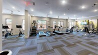 The Lexington Dilworth Fitness Center in Charlotte, NC offers a comprehensive workout facility spread across a single, open floor. The space is thoughtfully organized into distinct exercise zones, providing members with a well-rounded fitness experience. The cardio section features multiple treadmills, stair climbers, and upright bikes, all positioned along windowed walls that allow natural light to filter in and offer views of the surrounding brick exterior courtyard. A dedicated cycling area includes several stationary spin bikes arranged in front of floor-to-ceiling mirrors, which are complemented by a large mounted flat-screen TV, creating an immersive studio atmosphere. The strength training zone is equipped with a Smith machine, cable systems, weight racks, and various resistance machines, alongside wall-mounted TVs and a nature-inspired mural that adds a welcoming visual element. A functional training zone rounds out the space, outfitted with kettlebells, resistance bands, stability balls, and a pull-up bar station. Throughout the facility, patterned carpet tiles provide both comfort and visual interest, while recessed lighting and ceiling fans ensure a bright, comfortable environment. The overall ambiance is modern, clean, and purpose-driven.