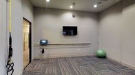 This single-floor fitness and wellness studio is designed with functionality and simplicity in mind. The space features neutral grey walls and textured carpet flooring that lends a clean, professional aesthetic throughout. A wall-mounted flat-screen television serves as a central focal point, ideal for guided workouts or instructional content. Below it runs a horizontal ballet barre fixed to the wall, providing support for stretching, balance training, or dance-based exercises. A small touchscreen control panel or tablet is mounted at one end of the barre, suggesting integrated technology for managing workouts or room settings. Recessed ceiling lighting, complemented by a single pendant light, ensures the space is evenly and adequately illuminated. A stability ball stored in the corner adds to the room's versatility, accommodating a range of fitness activities. A door on the far wall provides additional access or connects to an adjacent area. Overall, the studio offers a well-appointed, multipurpose environment suited for individual or small-group exercise sessions.