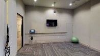 This single-floor fitness and wellness studio is designed with functionality and simplicity in mind. The space features neutral grey walls and textured carpet flooring that lends a clean, professional aesthetic throughout. A wall-mounted flat-screen television serves as a central focal point, ideal for guided workouts or instructional content. Below it runs a horizontal ballet barre fixed to the wall, providing support for stretching, balance training, or dance-based exercises. A small touchscreen control panel or tablet is mounted at one end of the barre, suggesting integrated technology for managing workouts or room settings. Recessed ceiling lighting, complemented by a single pendant light, ensures the space is evenly and adequately illuminated. A stability ball stored in the corner adds to the room's versatility, accommodating a range of fitness activities. A door on the far wall provides additional access or connects to an adjacent area. Overall, the studio offers a well-appointed, multipurpose environment suited for individual or small-group exercise sessions.