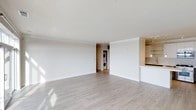 This single-floor space features an open-concept layout that maximizes its sense of spaciousness. The main living area is finished with light-toned hardwood flooring that runs throughout, complementing the crisp white walls and white ceiling to create a clean, airy atmosphere. Natural light filters into the space, casting soft highlights across the floor and enhancing the overall brightness of the interior.

A partial view of the kitchen is visible along one side, where warm wood cabinetry, a light countertop, and stylish pendant lighting add a touch of warmth and character to the otherwise minimalist palette. The kitchen appears to be separated from the main living area by a partial wall or breakfast bar, maintaining an open feel while defining each functional zone.

Toward the back of the space, a dedicated laundry area houses a stacked washer and dryer unit, providing practical in-unit convenience without compromising the overall layout. The transition between rooms is framed by clean architectural details, including subtle crown molding along the ceiling line, reinforcing the polished and well-finished character of this space.