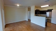 This single-floor space offers an open and functional layout that seamlessly connects the living area to the kitchen. The living area is finished with warm-toned hardwood flooring that extends throughout, lending a clean and inviting atmosphere to the space. Crown molding lines the ceiling, adding a subtle architectural detail that enhances the overall finish of the room. The kitchen is positioned adjacent to the living area, separated by a half-wall breakfast bar featuring granite countertops. Dark espresso cabinetry provides a striking contrast to the light walls and countertops, while a stainless steel microwave is integrated into the upper cabinet arrangement, contributing to a modern, polished look. The kitchen also benefits from dedicated overhead lighting, ensuring a well-lit workspace. A hallway visible from the main living area leads to additional rooms, suggesting a practical and well-organized floor plan. The neutral wall tones throughout create a versatile backdrop, allowing the space to adapt easily to various interior styles. Overall, the space presents a cohesive and well-maintained environment suitable for comfortable everyday living.