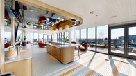 This generously sized single-floor amenity space delivers a striking blend of contemporary design and playful aesthetic sensibility. The interior is immediately distinguished by its signature mosaic tile flooring, rendered in a geometric pattern of gold, sky blue, and white — a cohesive visual thread that runs throughout the entire space.

Floor-to-ceiling glass curtain walls line much of the perimeter, inviting abundant natural light and framing panoramic city views. Multiple sets of glass doors open directly onto an outdoor terrace, seamlessly connecting interior and exterior living. A sculptural brass circular ceiling feature anchors the central kitchen and bar area, where a wood-paneled island with pendant lighting serves as both a functional and design focal point.

The layout accommodates a range of uses: a formal conference zone features a marble oval table surrounded by terracotta velvet chairs beneath a brass chandelier, while separate lounge areas offer curated seating with mid-century-inspired armchairs and accent rugs. Warm wood ceiling panels add texture overhead, and carefully placed indoor plants contribute a fresh, organic element. Curated artwork adorns select walls, completing the sophisticated, gallery-like atmosphere throughout.