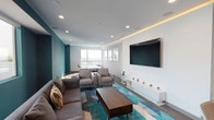 This single-floor amenity lounge is designed with a contemporary aesthetic, combining bold teal accent walls with crisp white ceilings and smooth hardwood-style flooring throughout. Recessed lighting, complemented by subtle warm cove lighting along the ceiling, ensures the space is well-illuminated while maintaining an inviting ambiance.

The main lounge area is anchored by a generous sectional sofa and pair of armchairs arranged around a solid wood coffee table, all set atop a geometric teal-toned area rug. A large wall-mounted flat-screen television is positioned above a sleek white media console, creating a dedicated entertainment zone. Adjacent to the seating area, a kitchenette offers counter seating, modern cabinetry with wood accents, and stainless steel appliances, making the space functional for gatherings.

Toward the far end of the lounge, a dedicated ping-pong area is bathed in natural light from expansive floor-to-ceiling windows that frame panoramic city views, including a visible urban skyline. These large windows extend along one wall, allowing abundant daylight to flood the interior throughout the day. The overall design balances recreational functionality with a polished, modern atmosphere well-suited for social and leisure activities.