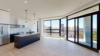 This contemporary single-floor residence is defined by its striking curved wall of floor-to-ceiling black-framed glass, which wraps around the open-plan living and kitchen area, flooding the interior with natural light and providing panoramic views of the surrounding urban skyline. A private balcony is accessible directly from the living space through full-height glass doors, extending the connection to the outdoors.

The kitchen is thoughtfully designed with two-tone cabinetry — white upper cabinets contrasted by dark lower units — complemented by a white grid-tile backsplash, stainless steel appliances including a range and built-in microwave, and a large dark island with a light countertop and matte black fixtures. Pendant lighting adds a refined touch above the island.

Light wood-look flooring runs throughout the living areas and bedrooms, contributing to the bright, airy atmosphere. Bedrooms feature large black-framed windows with city views and access to walk-in closets.

Two bathrooms are finished in a modern palette: one includes a soaking tub, dark vanity cabinetry, large-format grey tile flooring, and a wood-framed mirror, while the second features white subway tile, an open shower, and dark stone-effect floor tiles with matte black hardware throughout.