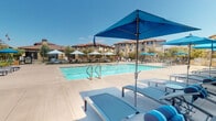 Located at 1692 Sycamore Dr in Simi Valley, California, this single-level outdoor amenity area offers a well-appointed resort-style experience within a Mediterranean-influenced residential community. The centerpiece is a generously sized swimming pool with crystal-clear blue water, flanked by a spacious concrete pool deck designed for both relaxation and socializing. Numerous cushioned chaise lounges with coordinating blue umbrellas are arranged poolside, providing ample shaded areas to unwind under the Southern California sun. Beyond the pool deck, an outdoor dining area features sturdy tables and chairs beneath additional shade umbrellas, creating an inviting space for al fresco gatherings. The perimeter is enclosed by a black wrought-iron fence, with young trees and landscaping adding natural greenery to the setting. A dedicated restroom facility, ADA-accessible for both men and women, is conveniently located on-site and includes a dual-level water fountain with a bottle-filling station. The surrounding residential buildings display classic Spanish-style architecture with red tile roofs and warm stucco exteriors, complementing the outdoor space's cohesive aesthetic. Under clear blue skies, this amenity area captures the essence of comfortable, community-centered California living.