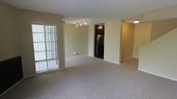 This two-floor space features a bright, open-concept main living area finished with neutral beige walls and plush wall-to-wall carpeting throughout. A large sliding glass door floods the room with natural light while providing direct access to an outdoor balcony or patio area, enhancing the sense of openness. Track lighting is mounted along the ceiling, offering adjustable illumination across the living space.

Adjacent to the main living area, the kitchen is distinguished by dark espresso-toned cabinetry and a stainless steel refrigerator, creating a modern contrast against the lighter surrounding walls. The transition between the living and kitchen areas is defined by a subtle architectural division, maintaining an open feel while clearly delineating the two functional spaces.

White baseboards provide clean, crisp framing throughout the room, contributing to a tidy and well-maintained aesthetic. The overall design is straightforward and functional, emphasizing a neutral, versatile palette that accommodates a variety of interior styles. With two floors, the space offers ample room for both living and private areas across separate levels.