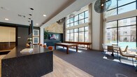This contemporary amenity lounge is designed to impress, combining sophisticated finishes with an open, social atmosphere. The space is anchored by dramatic double-height ceilings adorned with sculptural woven pendant lights, creating an immediate sense of grandeur upon entry. The entrance foyer features light wood-look flooring and sleek black-framed glass doors, setting a modern tone from the start.

The main lounge area transitions between hardwood-style flooring and plush carpeting, defining distinct activity zones. A striking natural wood accent wall with a herringbone pattern serves as a focal point, complemented by a wall-mounted flat-screen TV and curated artwork. Floor-to-ceiling windows line an entire wall, flooding the space with abundant natural light and offering direct views of an outdoor courtyard and resort-style pool flanked by palm trees.

The game area is well-equipped with a full-size pool table and a shuffleboard table, providing entertainment options for residents. A bar-height counter with leather-upholstered seating offers a casual dining and social setting. Comfortable lounge chairs and sofas are arranged throughout for relaxed conversation or casual gatherings. A cozy nook near the oversized windows features a small game table and seating, ideal for enjoying the outdoor views. The overall ambiance is polished, inviting, and thoughtfully curated.