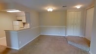 This single-floor residence offers a straightforward and functional layout designed for comfortable everyday living. The main living area is finished with wall-to-wall beige carpeting that adds warmth underfoot, while neutral-toned walls create a clean, versatile backdrop suited to a variety of décor styles. A decorative chair rail runs along the perimeter of the room, adding a subtle architectural detail that breaks up the wall space with a classic touch. Overhead, a flush-mount ceiling light fixture provides central illumination, and a ceiling vent ensures consistent climate control throughout the space. A bi-fold closet door along one wall offers convenient built-in storage. The living area opens directly to the kitchen via a pass-through counter, allowing for easy interaction between the two spaces. The kitchen is equipped with white cabinetry, a range, and a refrigerator, providing all the essentials in a compact and efficient arrangement. The open sightlines between the kitchen and living area give the home an inclusive, connected feel, making it well-suited for both daily routines and casual entertaining. Overall, the space presents a clean, move-in ready environment with practical finishes and a cohesive neutral palette throughout.