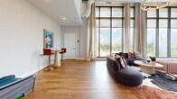 Located at 222 East Boyd Drive in Baton Rouge, LA, this 680-square-foot amenity lounge is a single-floor communal space designed with a modern industrial aesthetic. The room immediately impresses with its soaring ceilings, exposed metal ductwork, and an expansive wall of floor-to-ceiling steel-framed windows that flood the interior with natural light and offer sweeping outdoor views of the surrounding greenery and streetscape. Warm-toned hardwood flooring runs throughout the entire space, complementing the neutral gray walls and creating a cohesive, inviting atmosphere. A central seating area features a curved, contemporary sectional sofa paired with accent chairs and a coffee table, ideal for social gatherings. A full-sized pool table with a blue felt surface anchors the entertainment zone and is highlighted by a modern circular chandelier overhead. Flowing floor-length drapes frame the windows, softening the industrial elements. A compact bistro table with red bar chairs provides an intimate dining or casual work nook. A dedicated game alcove features a bold, graphic-patterned accent wall alongside a classic skee-ball arcade machine, adding a playful touch to the space. Together, these elements create a dynamic, amenity-rich environment suited for both relaxed socializing and active recreation.