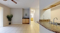 This single-floor residence is designed with a functional, open layout that connects its primary living spaces seamlessly. The main living area is finished with large-format ceramic tile flooring and features a ceiling fan overhead, a textured ceiling, and a built-in cabinet with a dark stone countertop and a display shelf above — a distinctive architectural detail that adds character to the space. Sliding glass doors fitted with vertical blinds allow abundant natural light and provide direct access to an outdoor area with views of surrounding greenery.

The kitchen is configured as a galley-style space with light wood cabinetry, dark stone-patterned countertops, a stainless steel sink, and white appliances including a refrigerator, range, and microwave. A pass-through window with a breakfast bar counter connects the kitchen directly to the living and dining area, enhancing the open feel.

The bedrooms are carpeted and each includes a ceiling fan. One bedroom features a walk-in closet and direct access to a bathroom and an in-unit laundry area with a stacked washer and dryer. The bathroom includes a wood-toned vanity with a stone-style countertop, dual mirrors, and a shower-tub combination with tile surround.