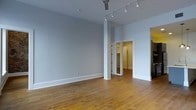 This single-floor space offers a clean, open layout defined by warm-toned hardwood floors and neutral light gray walls that create a bright, airy atmosphere. The main area is generously proportioned, with tall ceilings that enhance the sense of openness. A structural column adds architectural character while subtly dividing the space. Overhead, a track lighting system runs along the ceiling, providing flexible, adjustable illumination throughout the room, complemented by a ceiling fan for added comfort. Clean white baseboards frame the perimeter of the room, adding a crisp, finished look. Toward the rear of the main area, an open doorway leads into an adjoining room, which features mirrored closet doors that reflect light and contribute to a sense of added depth and space. The overall aesthetic is minimal and versatile, making the layout well-suited to a variety of uses. The combination of natural wood flooring, neutral tones, and purposeful lighting creates a cohesive, functional environment that is both welcoming and practical.