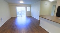 This single-floor space offers a clean, open layout designed for comfortable everyday living. The living area is a standout feature, showcasing rich, dark hardwood flooring that runs throughout and reflects the abundant natural light that pours in through a large sliding glass door fitted with vertical blinds. The door opens to a private balcony, providing both an outdoor retreat and a bright, airy atmosphere within. A secondary window further enhances the brightness of the space, keeping the interior feeling open and welcoming. The walls are finished in a neutral, warm white tone that complements the warm tones of the flooring. A half-wall partition with a wooden countertop edge subtly defines the transition between the living area and an adjacent space, adding architectural interest while maintaining the open feel. The overall ambiance is modern and understated, with a neutral palette that provides a versatile backdrop for a variety of interior styles. The space is thoughtfully laid out to maximize usability, making it well-suited for both relaxed living and functional day-to-day activities.