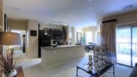 This single-floor residence showcases an open-concept layout that connects the kitchen, dining, and living areas into one cohesive, flowing space. The kitchen is a standout feature, equipped with dark espresso cabinetry that extends to the upper walls, black appliances including a refrigerator, range, and microwave, and a granite-topped central island that provides additional prep and seating space. Track lighting runs along the ceiling, illuminating both the kitchen and the adjacent living areas effectively.

The transition from kitchen to dining is seamless, with the dining area benefiting from natural light streaming through a prominent window. The living area features plush carpeting that adds warmth and comfort underfoot, contrasting with the clean, neutral-toned walls throughout. Decorative accents, including candle holders on a dark coffee table, contribute to the space's polished aesthetic.

The overall ambiance is modern and practical, with a neutral color palette of warm beige walls and dark fixtures creating a balanced, inviting atmosphere. The open floor plan maximizes the sense of space, making the interior feel airy and well-connected throughout.