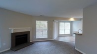 This single-floor space presents a well-designed open living area that combines comfort and functionality. The living room serves as the focal point, anchored by a dark-tiled fireplace with a clean white mantle, adding a classic touch to the neutral-toned interior. Soft, plush carpeting covers the floor, contributing to a warm and inviting atmosphere throughout the main living area.

Two generously sized windows dressed with horizontal blinds allow ample natural light to fill the room, offering views of the surrounding greenery outside. The windows create a bright, airy feel while maintaining privacy when needed. The transition from carpet to what appears to be hard-surface flooring near the adjacent area hints at a functional layout that separates living and utility zones.

The walls are finished in a soft, neutral beige tone, complementing the light-colored trim and baseboards throughout. The ceiling is smooth and standard in height, maintaining a clean, uncluttered look. The open sightlines between the living area and the adjoining space suggest a practical, connected floor plan suited for everyday living.