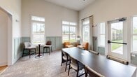 Located at 1120 Corner Wind Ln in Columbia, SC, this single-floor dining room is a thoughtfully designed gathering space that balances sophistication with comfort. The room immediately impresses with its soaring ceilings, which lend a sense of grandeur and openness well beyond its intimate footprint. Soft, neutral wall tones are complemented by elegant sage-green wainscoting that lines the lower walls throughout, adding a refined architectural detail that ties the space together.

Natural light pours in generously through multiple large double-hung windows dressed with sheer white curtains, offering pleasant views of the surrounding greenery. The flooring features a warm, textured area rug that anchors the varied seating arrangements. A central dark-wood conference-style table with upholstered chairs provides a flexible setting for group dining or meetings, while cozy upholstered booth seating along the walls creates more intimate nooks ideal for smaller gatherings.

A pair of warm-toned accent chairs and a bistro-style pedestal table occupy a sun-drenched corner, perfect for casual conversation. The walls are adorned with a striking gallery of dark, organically shaped wood art pieces alongside a large-scale landscape painting, adding an artful, curated character to the room. Pendant lighting with a modern touch completes the ambiance, making this dining space as visually engaging as it is functional.