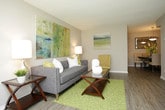 This single-floor residence offers a thoughtfully designed open-concept layout that seamlessly connects the living and dining areas, creating a cohesive and functional flow throughout the space. The living area features smooth wood-style flooring that extends throughout, complemented by soft neutral wall tones that lend a clean, welcoming ambiance. A textured ceiling adds subtle architectural character overhead. The living room is well-proportioned, providing ample space for seating arrangements, and is brightened by warm ambient lighting from a classic table lamp. Decorative wall art in muted, nature-inspired tones adds a layer of visual interest to the space. Transitioning naturally from the living area, the dining space is distinguished by an elegant pendant chandelier that anchors the room and casts a warm glow over the dining table. The dining area also features a decorative wall mirror and floral accents, adding a refined touch. The consistent flooring and coordinated color palette throughout both areas reinforce the open, unified feel of the interior. Overall, the space presents a comfortable and well-appointed single-level layout with attention to detail in both design and functionality.