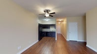 This single-story residence at Vineland Carriage Homes offers an open-concept living area that flows seamlessly into the kitchen, creating a cohesive and functional layout. The living space is finished with warm hardwood-style flooring that extends throughout, complemented by crisp white baseboards and soft, neutral-toned walls that lend a clean, welcoming ambiance. A ceiling fan with integrated lighting is centrally positioned overhead, providing both comfort and illumination. The textured ceiling adds a subtle design detail to the space. The kitchen is defined by dark espresso-toned cabinetry that contrasts elegantly with the light countertops and neutral walls. Black appliances, including a refrigerator, are built into the cabinetry layout, maintaining a cohesive color palette. A hallway visible from the main living area leads further into the home, suggesting additional rooms and providing a sense of depth. A white paneled door on the right side of the living space offers convenient access to an adjacent room or closet. The overall design is practical and cohesive, with a consistent flooring material tying each visible area of the home together.