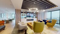 Located at 4607 N Broadway Street in Chicago's Uptown neighborhood, this thoughtfully designed amenity lounge offers a dynamic, single-floor social space that blends comfort and contemporary style. The layout is organized into several distinct zones, each with its own character and purpose.

At the heart of the space, a sleek built-in fireplace clad in marble-look stone anchors a central entertainment wall featuring a large flat-screen TV and flanking wood shelving. Overhead, a striking slatted ceiling detail with globe string lights adds warmth and visual interest. Multiple seating vignettes — including soft lounge chairs in muted and bold tones alongside low wooden coffee tables — create inviting areas for gathering or relaxing.

A dedicated wet bar area offers counter seating with orange upholstered barstools, patterned tile detailing, wall-mounted TVs, and floor-to-ceiling windows that frame expansive views of the Chicago skyline. Natural light pours in generously throughout.

Toward the perimeter, a dining nook features a round marble-top table and sage green chairs set against a textured, wave-patterned accent wall adorned with a curated gallery of framed artwork. Additional casual seating areas with hardwood flooring provide flexible space for games or conversation. The overall aesthetic is modern, curated, and welcoming.