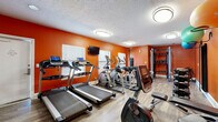 This single-level fitness center is defined by its energetic design, featuring bold orange walls and wood-style vinyl flooring throughout. The space is thoughtfully organized into distinct workout zones to accommodate a variety of training styles. The cardio area is outfitted with multiple treadmills and elliptical machines, each equipped with individual monitors, alongside a recumbent stationary bike. Natural light filters in through several windows, complementing the flush-mounted and recessed ceiling lighting overhead. A wall-mounted television provides entertainment during workouts. A full-wall mirror runs along one side of the room, visually expanding the space and allowing users to monitor their form. The strength training section features a multi-functional cable machine, a dumbbell rack with a varied weight selection, medicine balls, and a punching bag for boxing-style training. Wall-mounted brackets hold stability balls and a fold-down exercise mat, offering convenient access for stretching and core work. The textured ceiling adds a traditional finish to the space. Two separate entry doors provide easy access. Overall, the fitness center offers a comprehensive and energetic environment suitable for cardio, strength, and functional training.