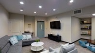 This single-floor space offers a thoughtfully designed communal lounge area that balances functionality with modern aesthetics. The room features smooth, light-toned walls paired with neutral gray carpeting, creating a clean and cohesive visual foundation throughout.

Recessed ceiling lights are evenly distributed across the space, providing bright, uniform illumination that enhances the open and welcoming atmosphere. A large wall-mounted flat-screen television serves as a central focal point, positioned above a sleek black media console that adds a contemporary touch.

The seating arrangement consists of multiple gray upholstered sofas and chairs, accented with teal and white throw pillows that introduce subtle pops of color. A round white coffee table anchors the seating area, contributing to the room's modern, minimalist character.

A teal-painted entry door with a narrow glass panel adds a distinct architectural accent and connects the lounge to adjacent areas. A small countertop or service ledge is visible near the entrance, suggesting convenient access to additional amenities.

Overall, the space presents a polished, well-appointed environment suited for socializing and leisure, with a design sensibility that is both practical and visually cohesive.
