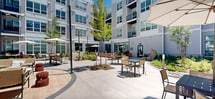 Located at 6 Becker Farm Rd in Roseland, NJ, this outdoor amenity space is a thoughtfully designed courtyard serving a modern multi-story residential community. The grounds are framed by sleek, contemporary buildings featuring large windows, horizontal siding, and private balconies that overlook the shared outdoor space below.

At the heart of the courtyard lies a resort-style swimming pool enclosed by black iron fencing, surrounded by lounge chairs and multiple market umbrellas. Curved, wide-format concrete walkways wind throughout the space, connecting distinct zones designed for both relaxation and socializing. Seating areas are furnished with a mix of casual outdoor furniture, including wicker egg chairs, colorful metal chairs, and dining-height table sets with umbrellas, offering residents a variety of settings to gather or unwind.

Lush landscaping features manicured lawns, mulched garden beds, ornamental shrubs, and young trees that provide greenery and shade throughout the space. A dedicated lawn area accommodates recreational games such as cornhole, while string lights overhead add ambiance for evening use. The overall design balances modern aesthetics with functional, community-oriented outdoor living in a vibrant New Jersey setting.