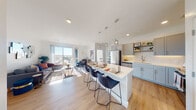This well-appointed single-floor residence showcases a thoughtfully designed open-concept layout that seamlessly connects the kitchen, dining, and living areas. The kitchen is a standout feature, outfitted with soft gray shaker cabinets, white subway tile backsplash, stainless steel appliances — including a refrigerator, range, microwave, and dishwasher — and open shelving for added storage and display. A large central island with a white quartz-style countertop and seating for multiple bar-height chairs serves as both a functional workspace and a casual dining hub, illuminated by a trio of modern pendant lights overhead. Light wood-style flooring flows throughout the main living areas, enhancing the bright and airy atmosphere. The living room is generously sized and bathed in natural light from floor-to-ceiling sliding glass doors that open to a private balcony, offering elevated outdoor views of the surrounding cityscape. The two bedrooms are carpeted for comfort and each features a distinctive accent wall — one with a textured script-style wallcovering and the other with a soft neutral textile-look finish. The bathroom includes a full soaking tub with a white tile surround and brushed gold fixtures. Recessed lighting and clean white walls throughout contribute to the space's modern, cohesive aesthetic.
