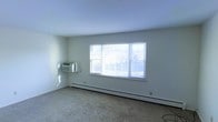 This single-floor space features a spacious main living area characterized by its clean, minimalist design. The room is finished with light gray carpet and smooth white walls, creating a neutral and versatile backdrop. A large, three-panel window serves as the focal point of the room, allowing abundant natural light to fill the space while offering views of the surrounding outdoor environment. Below the window runs a baseboard heating unit, providing efficient warmth throughout the room. A wall-mounted air conditioning unit on the adjacent wall ensures year-round climate control. The ceiling is smooth and painted white, contributing to the room's bright and airy atmosphere. The open layout of the living area provides flexibility for various furniture arrangements. Overall, the space presents a straightforward and functional environment, well-suited for comfortable everyday living.