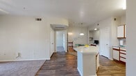 This single-floor residence offers a thoughtfully designed open-concept layout that connects the kitchen, dining, and living areas into one cohesive, light-filled space. The kitchen is well-equipped with ample white cabinetry accented by warm wood trim, a full suite of white appliances, a double sink, and generous counter space. A central island with a white countertop serves as the focal point of the space, providing additional workspace and a natural divide between the kitchen and living areas. Wood-style flooring runs throughout the main living spaces, lending a clean and consistent aesthetic. Adjacent to the kitchen, a dedicated dining area includes a built-in desk and open shelving — a practical feature ideal for a home workspace or display area. The bedroom is generously proportioned, featuring plush carpeting, a ceiling fan, and a large window that invites natural light and views of the surrounding greenery. The full bathroom includes a shower-tub combination and wood-style flooring that mirrors the rest of the unit. A curved architectural detail above the hallway entry adds a subtle design flourish to the otherwise clean, neutral interior. Overall, the space presents a functional and comfortable layout with a neutral palette suited to a variety of interior styles.