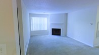 This single-story space centers around a living area that combines functional design with comfortable features. The room is finished with wall-to-wall carpeting in a neutral tone, providing a soft underfoot feel that extends throughout the space. A fireplace with a clean white mantel serves as a focal point along one wall, offering both visual interest and a practical heating element. Adjacent to the fireplace, a generously sized window fitted with vertical blinds allows for adjustable natural light, brightening the room while maintaining privacy. The walls are painted in a light, neutral tone, contributing to an open and airy atmosphere. A textured popcorn ceiling adds a classic finish overhead. The overall layout is open and unfurnished, allowing for flexible furniture arrangement to suit a variety of living styles. The neutral color palette throughout creates a cohesive and versatile backdrop, making the space easy to personalize.