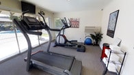 This single-floor fitness room offers a practical and inviting space for exercise and wellness. The room is outfitted with key cardio equipment, including a treadmill and an elliptical cross-trainer, providing users with versatile workout options. A stability ball and additional accessories are neatly arranged in the corner alongside a decorative potted plant, adding a touch of greenery to the space. Natural light filters in through a large window overlooking a landscaped outdoor area, creating a bright and energizing atmosphere. A wall-mounted air conditioning unit ensures year-round climate comfort during workouts. The room features neutral-toned carpeted flooring suited for a fitness environment, and the walls are kept clean and minimal, accented by cycling-themed artwork that complements the active theme of the space. A wall-mounted television is positioned for convenient viewing during exercise. The overall ambiance is functional and focused, making it an ideal space for residents or guests looking to maintain their fitness routine in a comfortable, well-maintained setting.