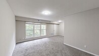 Located at 1301 Canal Dr in Chesapeake, VA, this single-floor, two-bedroom, one-bathroom home offers a practical and comfortable layout. The galley-style kitchen is a standout feature, equipped with slate-grey cabinetry, granite-look countertops, a double stainless steel sink, and white appliances including a range, microwave, and refrigerator. Warm wood-look flooring runs throughout the kitchen, adding a clean, modern contrast to the cabinetry. A hallway connects the main living areas and leads to the bedrooms and bathroom. The spacious primary bedroom offers generous square footage, soft carpeting, and ample natural light, while the second bedroom also features plush carpet and a window overlooking the greenery outside. Both rooms are finished with neutral, warm-toned walls and clean white trim, creating a calm and inviting atmosphere. The dedicated closet includes built-in shelving and hanging rods, providing well-organized storage. The bathroom is outfitted with a full tub-shower combo featuring classic white subway tile surround, a toilet, and a vanity mirror. Throughout the home, the consistent neutral palette and carpeted living spaces contribute to a cozy, move-in-ready feel.